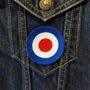 May include: A blue, white, and red target patch with a white circle in the center and a red circle inside the white circle. The patch is sewn onto a blue denim jacket.