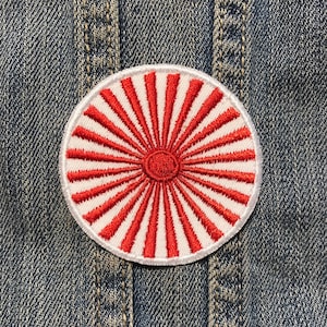 May include: Embroidered patch featuring a red sunburst design on a white background. The patch has a circular shape and is set against a denim background. The sunburst design radiates from a central red circle.