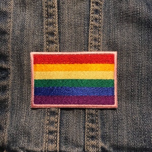 May include: A rainbow pride flag patch with red, orange, yellow, green, blue, and purple stripes. The patch is sewn onto a denim jacket.