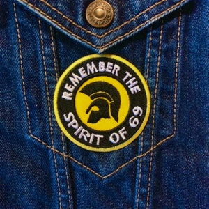 Op de afbeelding: Een geborduurde zwarte en gele patch met een witte rand. De patch heeft een Spartaanse helm en de tekst "Remember the Spirit of 69".