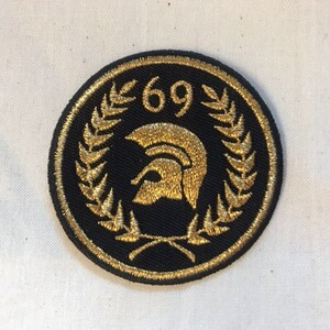 Puede incluir: Un parche bordado en negro y dorado con un casco espartano, una corona de laurel y el número 69.