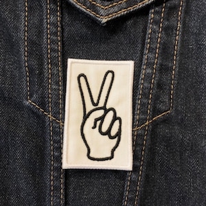 Può includere: Una toppa bianca a forma di segno della pace con contorno nero, cucita su una giacca di jeans scura.