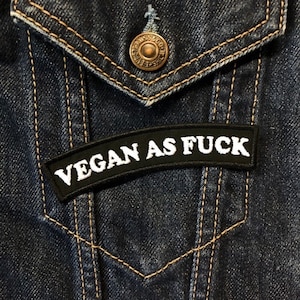 Könnte beinhalten: Ein schwarzer Aufnäher mit weiß gesticktem Text "VEGAN AS FUCK". Der Aufnäher ist auf einer blauen Jeansjacke angebracht. Die Jacke hat einen Knopfverschluss und sichtbare Nähte.
