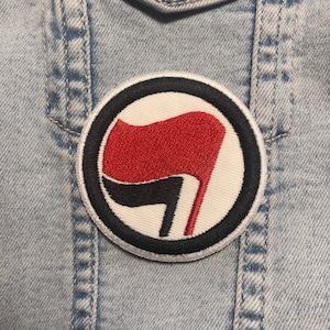 May include: A white, black, and red embroidered patch with a stylized fist holding a red flag. The patch is circular and has a black border.