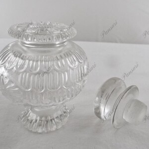May include: A clear glass bottle with a textured design and a matching stopper. The bottle has a round body and a wide base.