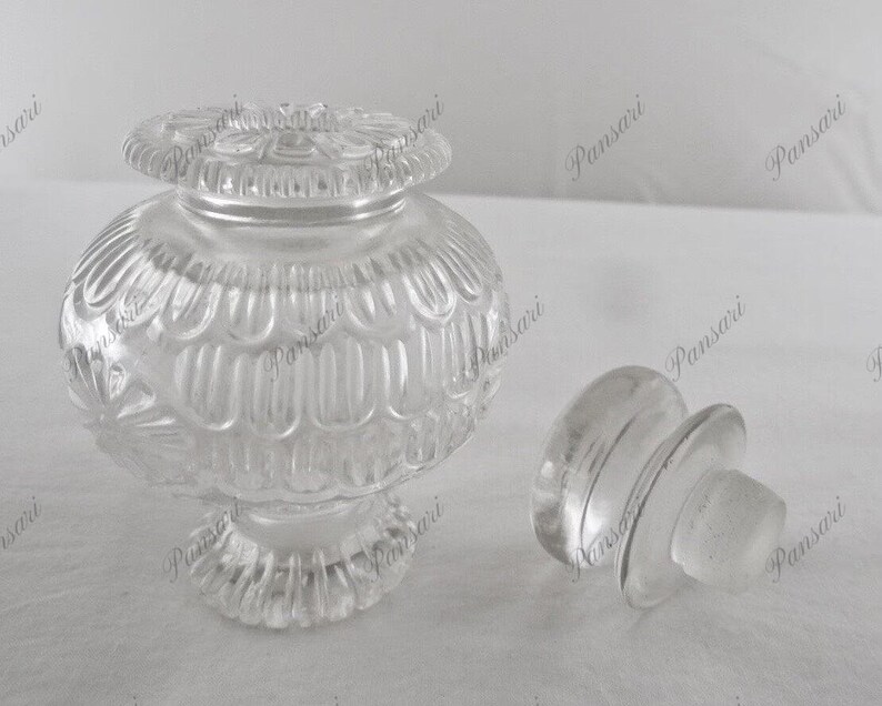 May include: A clear glass bottle with a textured design and a matching stopper. The bottle has a round body and a wide base.