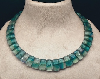 May include: A faceted, graduated necklace with a mix of teal and blue-green stones. The necklace is displayed on a neutral-toned mannequin bust. The stones are cut into rectangular shapes and are strung together to form a collar.