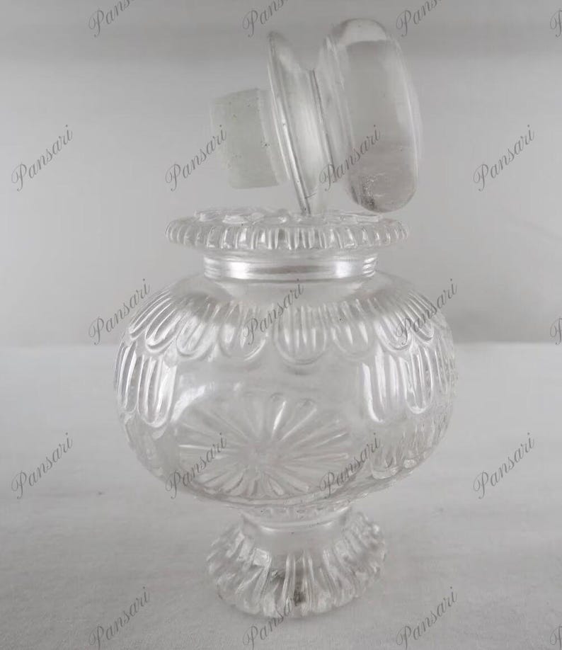 May include: A clear glass bottle with a textured surface and a decorative lid. The bottle has a round body and a wide base.