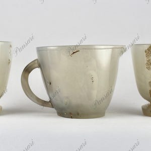 May include: Three antique cups made of white agate with brown markings. The cups are all different shapes and sizes. The cup on the left is a goblet, the cup in the middle is a pitcher, and the cup on the right is a smaller goblet.