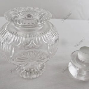 May include: A clear glass jar with a decorative lid. The jar has a round body with a textured design and a wide mouth. The lid has a similar design and fits snugly on top of the jar.