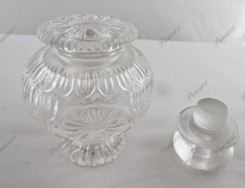 May include: A clear glass jar with a decorative lid. The jar has a round body with a textured design and a wide mouth. The lid has a similar design and fits snugly on top of the jar.