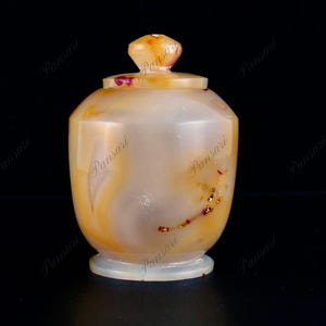 Puede incluir: Un pequeño tarro redondo de ágata con tapa. El tarro es de color naranja claro con remolinos marrones y dorados. La tapa es de color naranja oscuro.