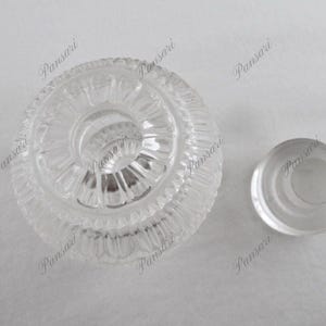 May include: A clear glass bottle stopper with a decorative, concentric circle design. The stopper has a smooth, rounded top and a flat base.