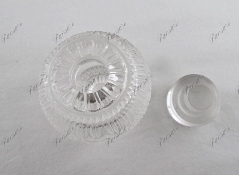 May include: A clear glass bottle stopper with a decorative, concentric circle design. The stopper has a smooth, rounded top and a flat base.