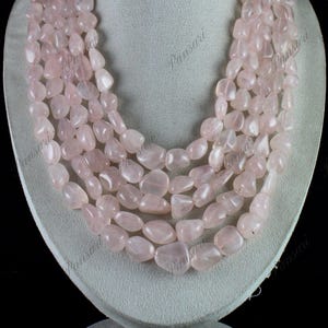 Puede incluir: Un collar de varias hebras hecho con cuentas de cuarzo rosa claro. Las cuentas son pulidas y tienen un acabado liso y brillante. El collar se muestra en un busto de maniquí blanco.