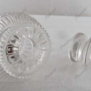 May include: A clear glass bottle stopper with a decorative, textured top. The stopper has a round, flat base.