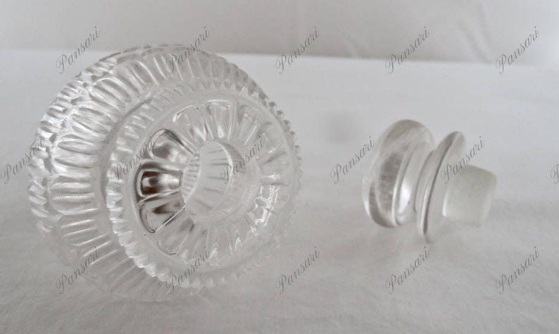 May include: A clear glass bottle stopper with a decorative, textured top. The stopper has a round, flat base.