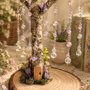 Puede incluir: Una escultura de &aacute;rbol decorativa sobre una base de rodaja de madera circular con una peque&ntilde;a puerta de madera en el tronco, musgo verde y detalles florales morados. Alambre plateado envuelve las ramas, adornadas con cuentas transparentes y rosas.