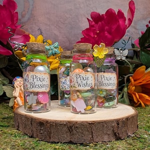 May include: Three small clear glass bottles with cork stoppers, filled with colorful beads and charms. Each bottle features a paper label reading Pixie Blessings or Pixie Wishes, decorated with wire butterflies, flowers, and small metal accents.