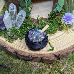May include: A miniature black cauldron filled with iridescent glass beads and a wooden stirring stick. The cauldron is sitting on a wooden slice with moss and crystals around it.