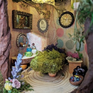 May include: A miniature fairy house with a wooden floor, a green moss bush, a small table with bottles and a mirror, and a clock on the wall. The house is decorated with flowers and vines.