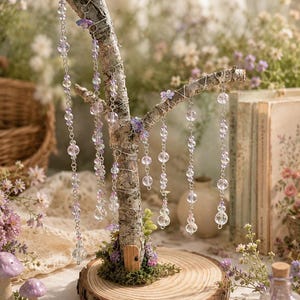 Puede incluir: Un soporte decorativo en forma de &aacute;rbol hecho de una rama natural con corteza, montado sobre una base circular de rodaja de madera. Varias cadenas plateadas con cuentas de vidrio facetado transparentes y moradas cuelgan de las ramas con flores.