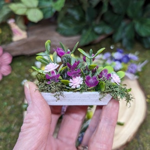 May include: A miniature rectangular white planter filled with artificial green foliage, small purple leaves, and tiny light pink flowers nestled in green moss. The planter has a speckled texture and is held in a palm to show its small scale.