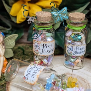May include: Three small glass bottles with cork stoppers, filled with colorful beads and charms. Two bottles stand upright, each featuring a label that reads Pixie Blessing and a wire butterfly accent. One bottle lies on its side, labeled Pixie Wishes.