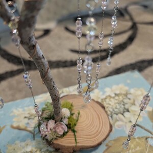 Puede incluir: Una escultura de &aacute;rbol decorativa con una base de rama de madera natural y peque&ntilde;os detalles florales en rosa y verde. Varias cadenas plateadas cuelgan de la rama, adornadas con cuentas de cristal facetadas transparentes, iridiscentes y rosa claro.