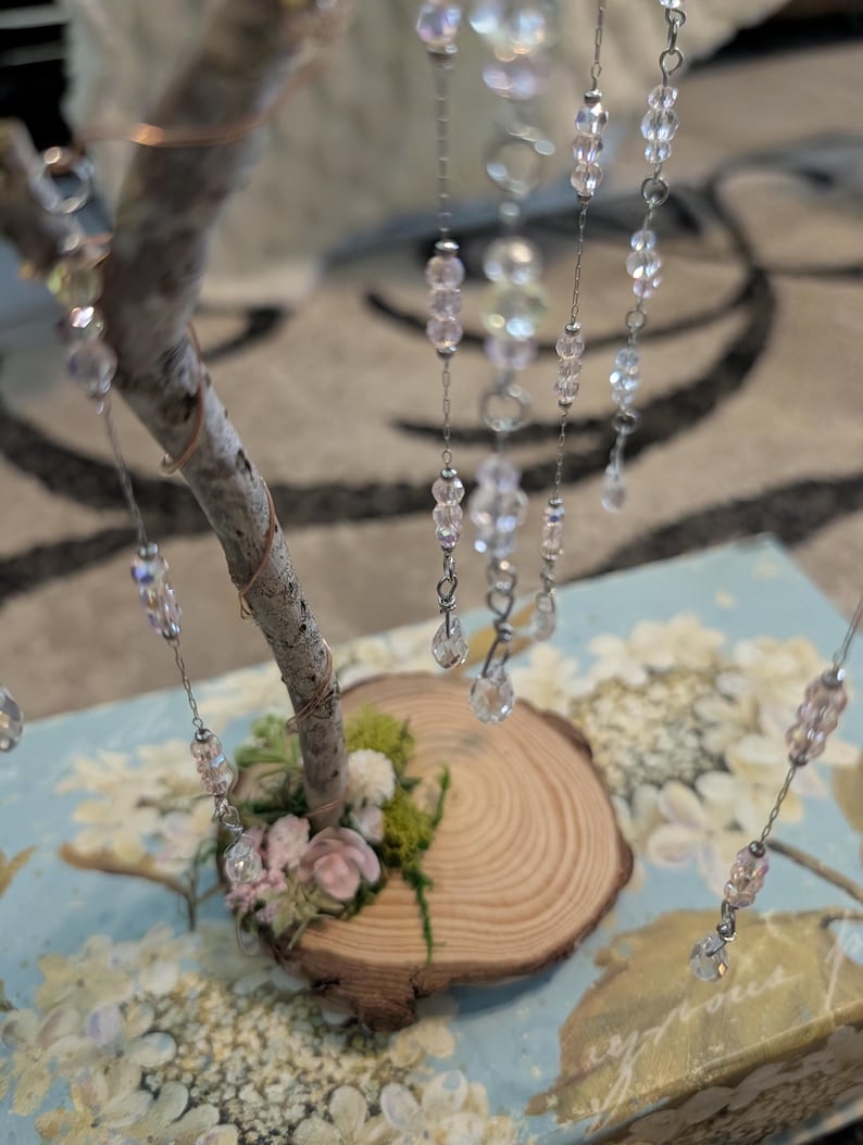 Puede incluir: Una escultura de &aacute;rbol decorativa con una base de rama de madera natural y peque&ntilde;os detalles florales en rosa y verde. Varias cadenas plateadas cuelgan de la rama, adornadas con cuentas de cristal facetadas transparentes, iridiscentes y rosa claro.