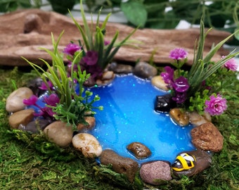 May include: A miniature fairy garden scene with a blue resin pond surrounded by small rocks and green plants. A small yellow and black bee figurine is sitting on a rock near the pond.