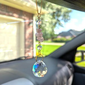 May include: A car charm featuring a faceted crystal pendant with a rainbow effect, suspended by a gold chain. The charm includes clear beads and pink rose quartz stones, creating a decorative and spiritual accessory.
