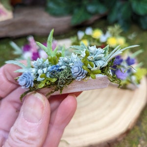 May include: A miniature wooden planter filled with faux succulents and flowers. The planter is painted white and has a rustic, weathered look. The succulents are a variety of shades of blue and green, and the flowers are pink and purple.