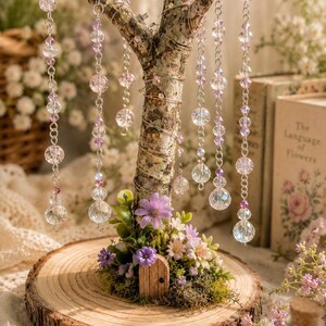 Puede incluir: Un &aacute;rbol decorativo sobre una base de rodaja de madera circular con una peque&ntilde;a puerta de madera, musgo verde y flores artificiales moradas y blancas. Varios hilos colgantes de cuentas de cristal facetadas transparentes y moradas.