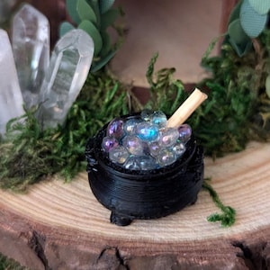 May include: A miniature black cauldron filled with iridescent glass beads and a wooden stirring stick. The cauldron sits on a wooden slice surrounded by moss, with clear quartz crystals and greenery in the background.