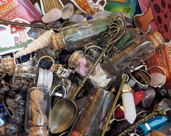 Op de afbeelding: Een verzameling kleine glazen flesjes gevuld met verschillende items, waaronder kristallen, gedroogde bloemen en andere natuurlijke elementen. De flesjes zijn gerangschikt in een houten doos met andere decoratieve items, zoals ringen, een lepel en een schelp.