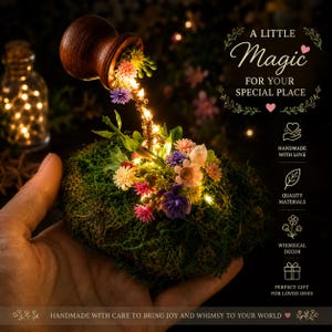 May include: A decorative miniature display featuring a small brown wooden pot pouring a stream of warm LED fairy lights onto a bed of green moss adorned with tiny colorful artificial flowers. Text reads A LITTLE MAGIC FOR YOUR SPECIAL PLACE.