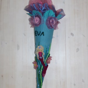 May include: A blue felt cone with a mermaid design, pink flowers, and the name "EVA" written on it. The cone is decorated with beads and sequins.