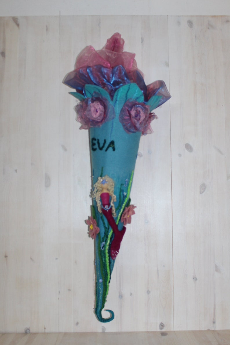 May include: A blue felt cone with a mermaid design, pink flowers, and the name "EVA" written on it. The cone is decorated with beads and sequins.