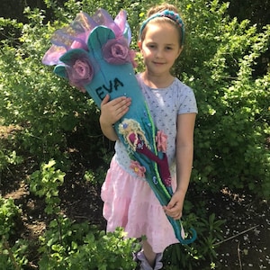 May include: A young girl holds a blue and purple felt school cone with the name "EVA" written on it. The cone is decorated with flowers and a mermaid illustration.