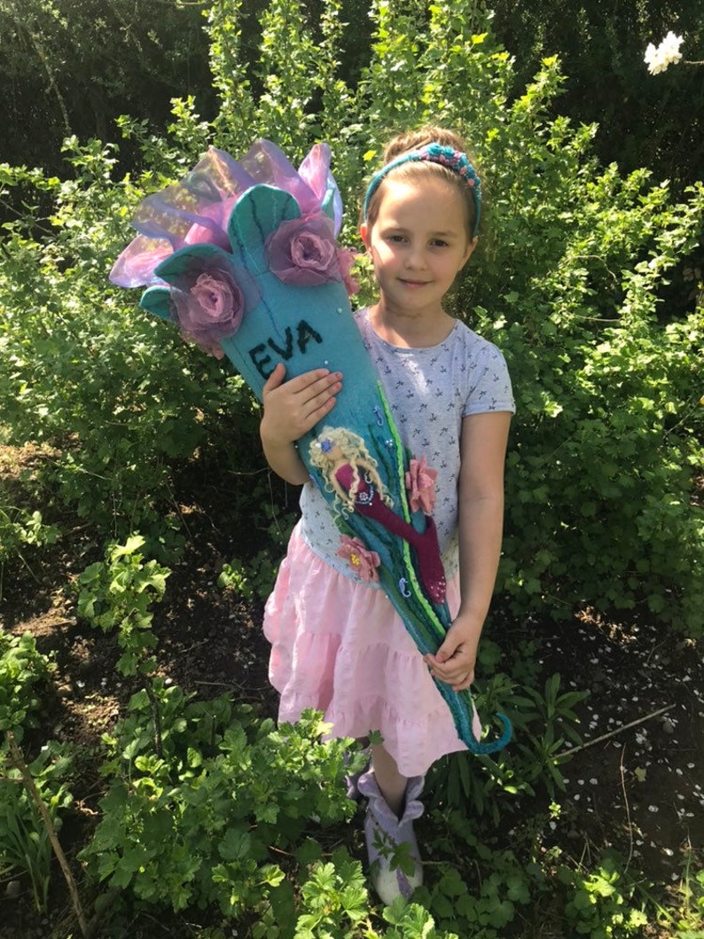 May include: A young girl holds a blue and purple felt school cone with the name "EVA" written on it. The cone is decorated with flowers and a mermaid illustration.