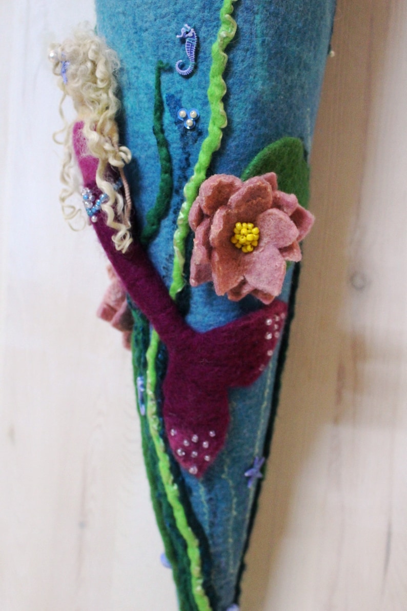 May include: A felt mermaid tail ornament with a pink flower, green seaweed, and a blue seahorse. The mermaid has long blonde hair and a pink tail.