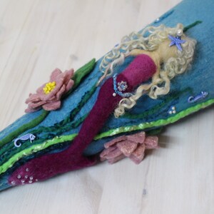 May include: A felt mermaid doll with long blonde hair, a pink tail, and a blue and green background. The mermaid is decorated with beads, sequins, and a starfish.