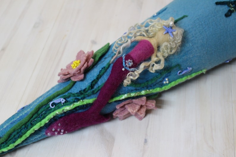 May include: A felt mermaid doll with long blonde hair, a pink tail, and a blue and green background. The mermaid is decorated with beads, sequins, and a starfish.