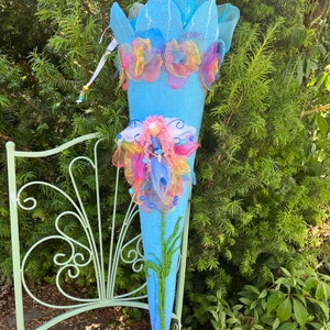 May include: A blue felt cone decorated with colorful flowers and a fairy figurine. The cone is sitting on a green metal chair.