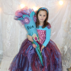 May include: A young girl in a purple and blue dress holds a blue and pink felt school bag with the name "EVA" embroidered on it.