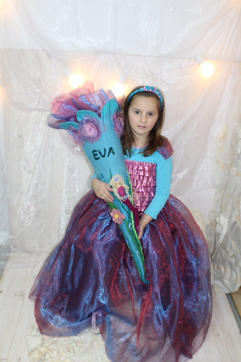 May include: A young girl in a purple and blue dress holds a blue and pink felt school bag with the name "EVA" embroidered on it.