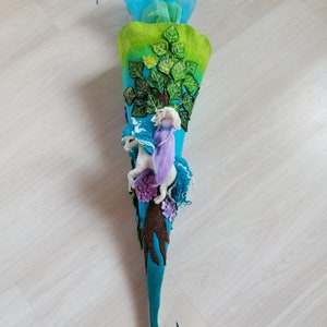 Könnte beinhalten: Eine handgefertigte Schultüte aus Filz in Türkis, Grün und Blau. Die Tüte ist mit einem Baum, einem Einhorn und einer Fee verziert. Der Baum hat grüne Blätter und braune Äste. Das Einhorn ist weiß und die Fee trägt ein lila Kleid.