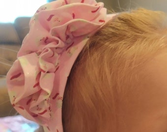 May include: A pink headband with a ruffled flower design. The headband has a white background with a pink and red floral pattern.