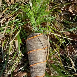 May include: A brown carrot-shaped object wrapped in twine with orange string accents. The object is sitting on a bed of green grass and brown leaves.